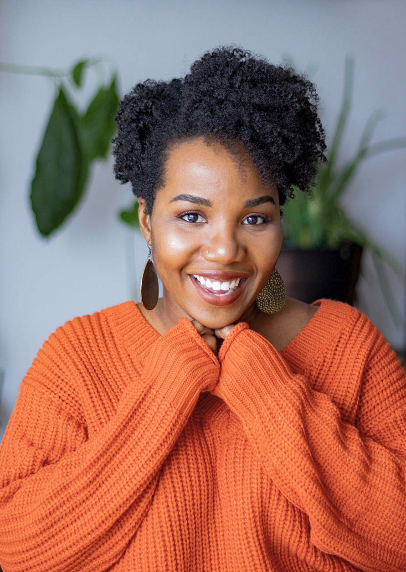 Portrait souriant d’Esther Nelsa en chandail orange, devant des plantes
